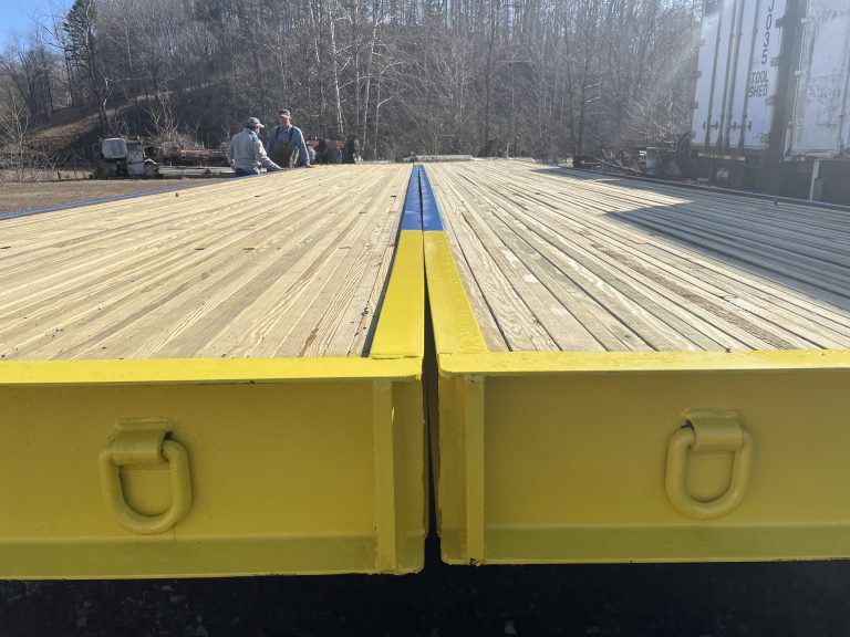 Timber bridge components with yellow edges and wooden planks, featuring workers in the background, highlighting E&H Manufacturing's specialty in stress-laminated timber bridge production.