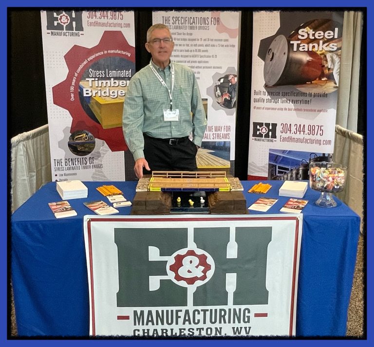 E&H Manufacturing representative at trade show booth showcasing stress-laminated timber bridge model and promotional materials, emphasizing products for the oil and natural gas industries.
