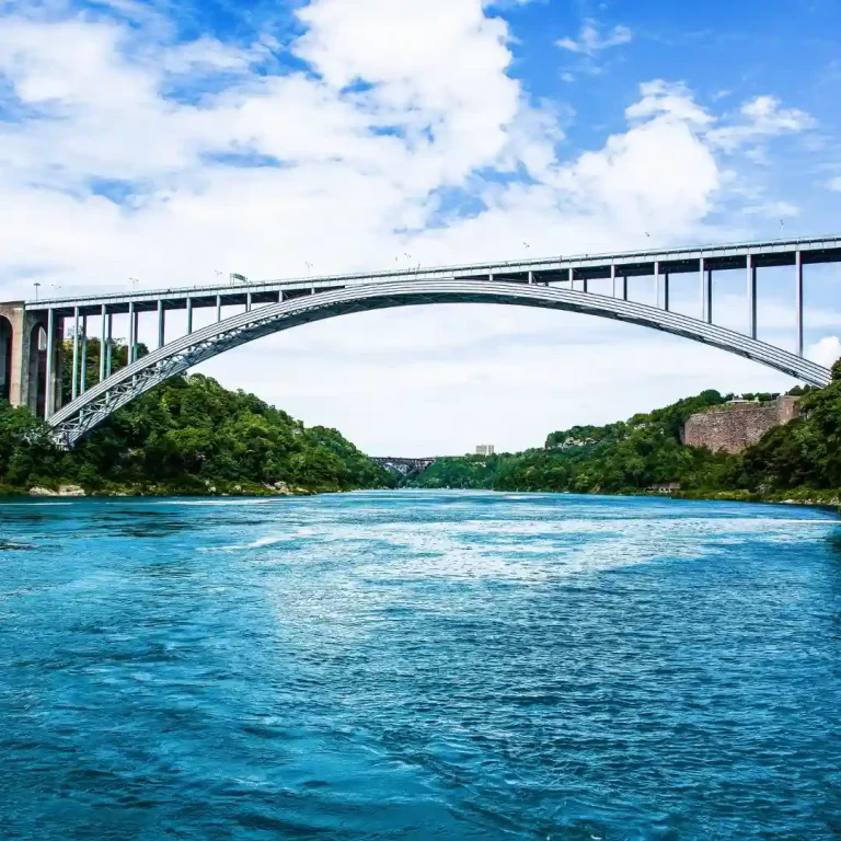 Steel arch bridge spanning a river, surrounded by lush greenery and a clear blue sky, representing temporary or permanent bridge solutions in infrastructure.