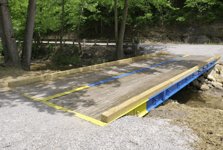 Stress-laminated timber bridge by E&H Manufacturing, featuring a wooden deck with blue and yellow accents, surrounded by greenery and gravel path.
