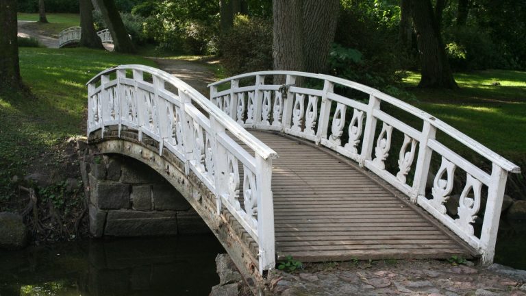 White timber bridge with decorative railings spanning a small stream in a lush green park, illustrating aesthetic and functional design principles relevant to timber bridge discussions.