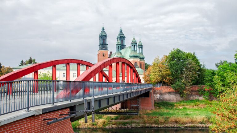 Red stress-laminated timber bridge spanning a river, with historical buildings and greenery in the background, illustrating innovative engineering in bridge design.