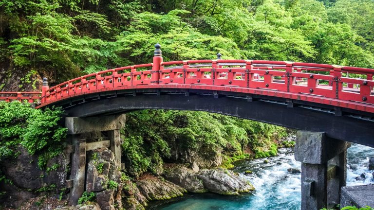 Red timber bridge arching over a flowing river, surrounded by lush greenery, illustrating the aesthetic and functional design principles of timber bridges.