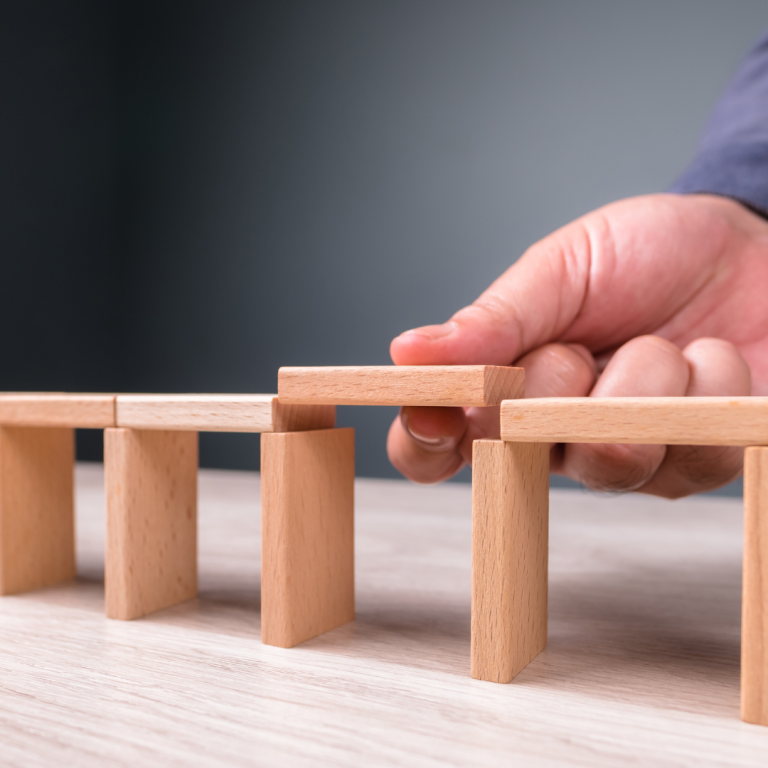 Hand placing a wooden block on top of two vertical wooden supports, symbolizing the construction of temporary bridges and accessibility solutions in engineering.