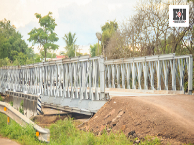Temporary metal bridge spanning a dirt road, showcasing E&H Manufacturing's portable infrastructure solutions, with lush greenery in the background.
